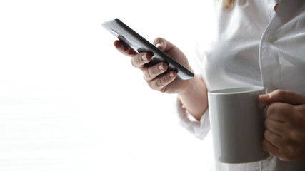 Young woman connected using a mobile taking a hot drink in a mug. White background and empty copy space for Editor's text.
