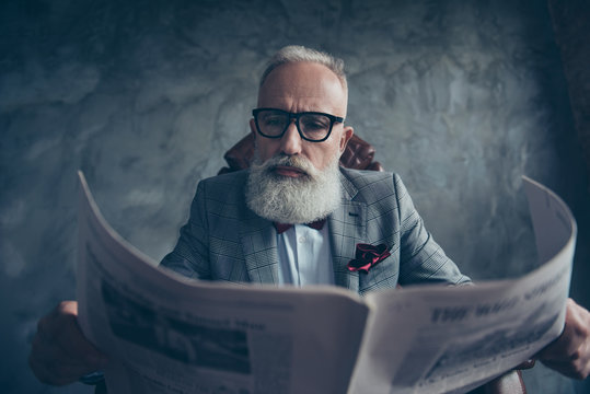 Attractive, smart, cool, old businessman in glasses and jacket reading newspaper, news about politic, bitcoin, finance, economy, science over gray background, sitting in chair