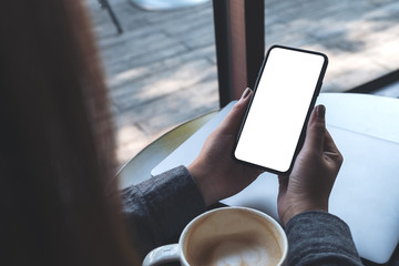 Mockup image of a woman holding black mobile phone with blank white desktop screen with coffee cup and laptop on table in cafe