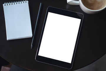Top view mockup image of a black tablet pc with blank white desktop screen with notebook and coffee cup on the table