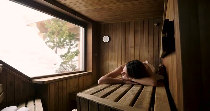 A beautiful woman wearing a white towel takes a sauna: The sauna is made of wood with a large window with a view of the snow. Concept of: relax, vacation, wellness center.