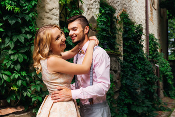 Fototapeta premium girl laughing and hugging her boyfriend in a pink shirt and wooden bow tie against a background building with bushes