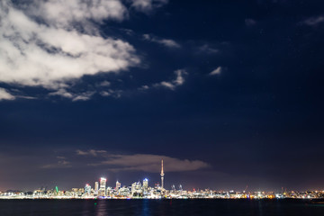 starry sky above Auckland city&rsquo;s skyline with some clouds and the harbour, the ocean and some boats at the front