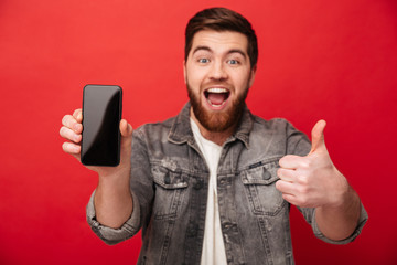 Portrait of unshaved cheerful guy demonstrating cell phone on camera and gesturing thumb up,...