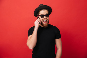 Portrait of irresistible fancy man 30s in sunglasses wearing black t-shirt and hat smiling and talking on mobile, isolated over red background