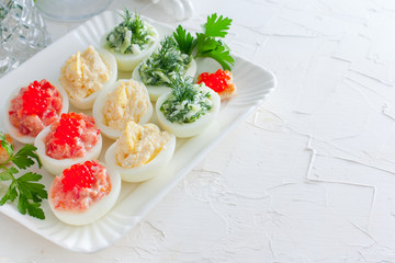 Festive decoration variants of stuffed eggs - red (red fish, caviar), yellow (cheese, eggs) and green (fresh cucumber, dill) on a white dish, horizontal, copy space