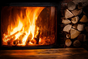 Old wooden table and fireplace with warm fire at the background. - obrazy, fototapety, plakaty