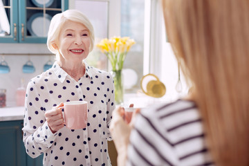 Delicious coffee. The focus being on a charming senior woman standing in the kitchen and drinking coffee together with her daughter and chatting with her