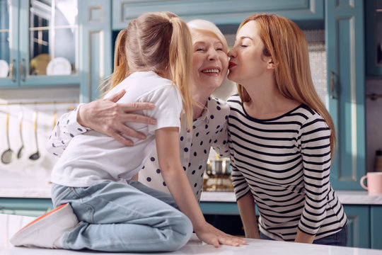 Tender kisses. Happy elderly woman standing in the kitchen and receiving kisses on the cheeks from her daughter and her little granddaughter sitting on the kitchen counter