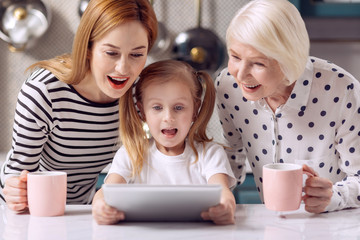 Exciting cartoon. Pleasant little girl watching a cartoon on tablet together with her grandmother and mother drinking coffee from pink mugs while looking amused by plot twists