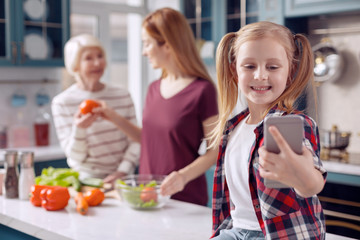 Peace at home. Pleasant young woman sitting on the kitchen counter and taking selfies of herself and her mother and grandmother cooking salad