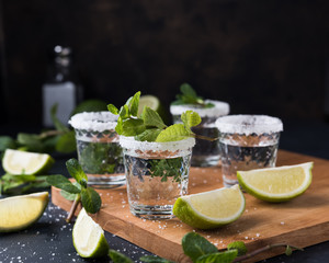 Alcohol shots on wooden tray. Shooter glasses of vodka or Tequila on black table. Mojito with mint