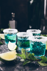 Alcohol shots on metal tray. Shooter glasses of blue vodka or Tequila on black table. Mojito with mint