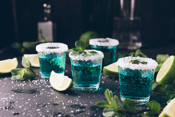 Alcohol shots on metal tray. Shooter glasses of blue vodka or Tequila on black table. Mojito with mint