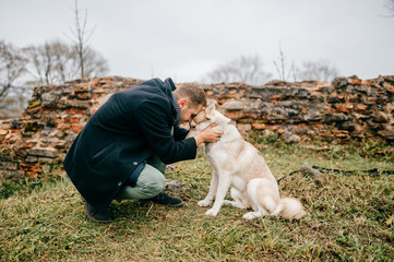 Adult handsome man in business clothes walking with cute lovely husky puppy dog outdoor beyond brick wall of european castle remains. Male owner with loving purebred canine hugs. Boy embracing his dog