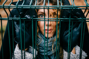 Odd bizarre fancy unusual frightened young girl closeup outdoor portrait. Adult female in jail. Expressive scared eyes look through steel wire. Comic funny emotional girl face hidden inside coat.