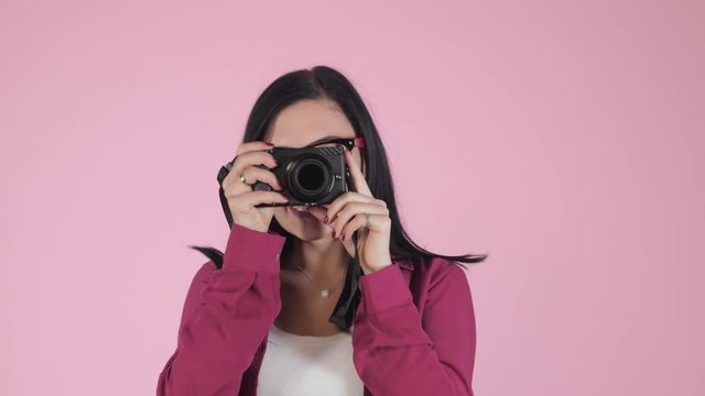 Young pretty brunette woman in pink shirt takes pictures with DSLR camera over colorful background in studio. Girl smiling, flirting and having fun as photographer. 4k