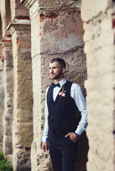 Fototapeta na wymiar Groom at wedding tuxedo smiling and waiting for bride near brick buildingl. Elegant groom in costume and bow-tie.