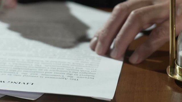 Old man reading and signing living will papers, sand flowing in hourglass table