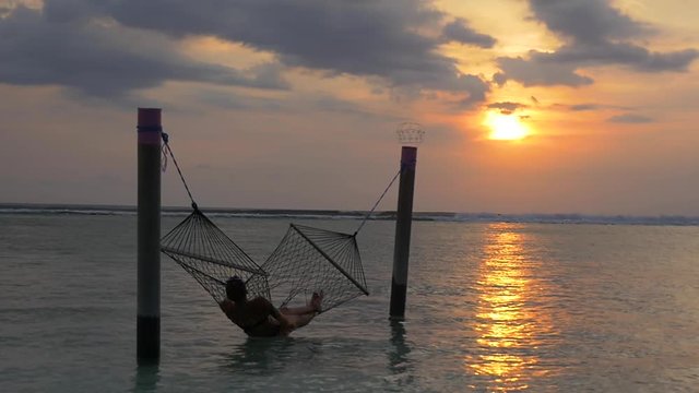 Waves and sunset backdrop. Silhouette of woman in a bikini relaxing in a hammock in the ocean