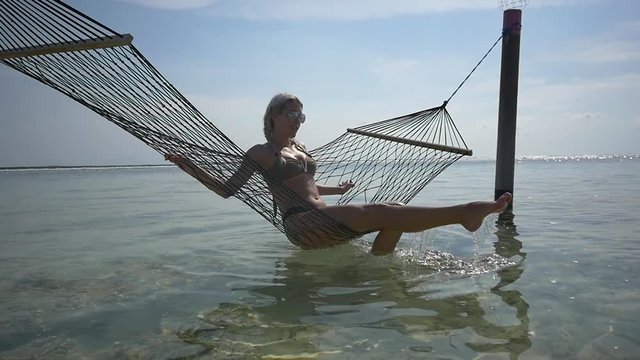Playful young woman in bikini sits and relaxes in a hammock in the ocean