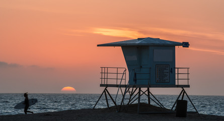 Naklejka premium Surfer biegnący obok wieży ratownika o zachodzie słońca na plaży Huntington w południowej Kalifornii