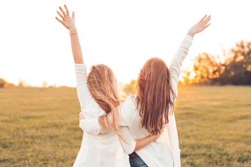 Two pretty girls raised their hands on a field at sunset. - obrazy, fototapety, plakaty