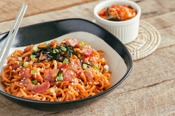 Korean hot and spicy instant noodle in black and white bowl topped with bacon, sesame, seaweed and chopped scallion served with kimchi on wood table. Homemade delicious asian food concept.