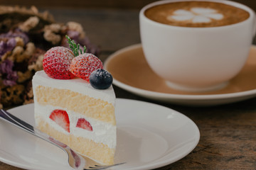 Homemade delicious and soft strawberry shortcake on wood table served with hot cappuccino coffee at bakery shop or cafe restaurant. Vanilla sponge cake with whipped cream and strawberries and icing.