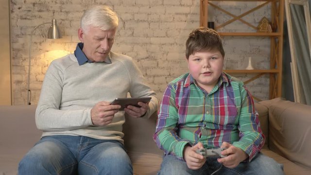 Grandfather and grandson are sitting on the couch. An old man uses a tablet, a young fat guy plays on the console game, loses. Video games. Home comfort, family idyll, cosiness concept, difference of