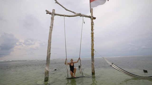 Beautiful young lady stands on a wooden swing in the ocean