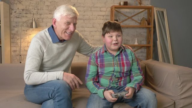 Grandpa enjoys the victory of his grandson in the console game. The old man is sitting on the couch with a young fat boy. Video game. Home comfort, family idyll, cosiness concept, difference of