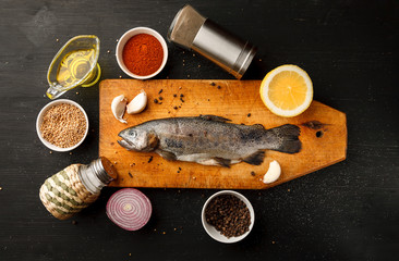 Fresh fish with herbs and spices on vintage cutting board on dark background, top view