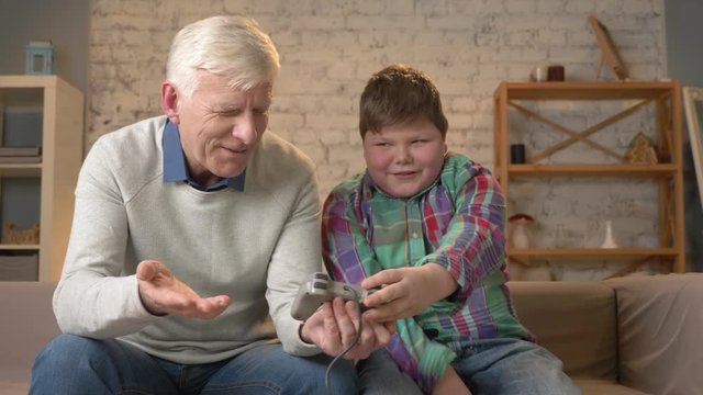The grandson teaches grandfather to play the console game. Elderly man is learning how to play a video game, he does not understand. Home comfort, family idyll, cosiness concept, difference of