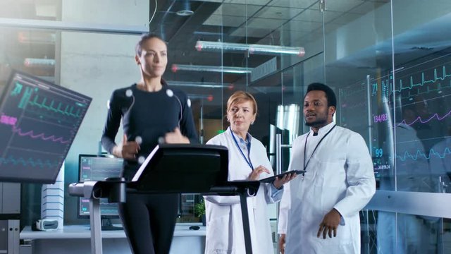 Woman Athlete Runs on a Treadmill  with Electrodes Attached to Her Body while Two Scientists Supervise whole Process. In the Background Laboratory with Monitors Showing EKG Readings.