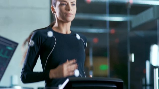 Beautiful Woman Athlete with Electrodes Connected to Her Body Runs on a Treadmill in a Sport Science Laboratory. In the Background High-Tech Laboratory with Monitors Showing EKG Readings.