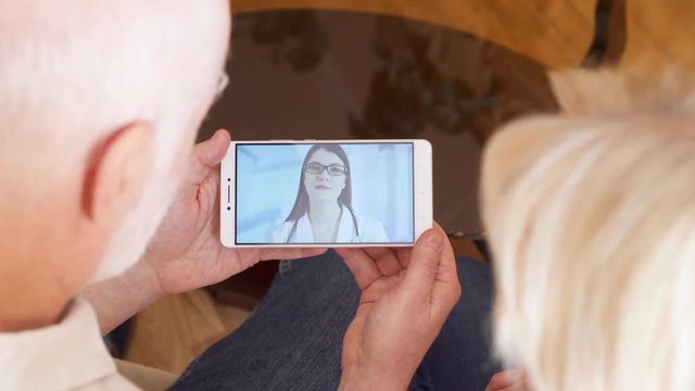 Top view of senior couple at home having medical consultation video chat on mobile with physician. Confident professional female doctor with stethoscope talking with patients via messenger app call