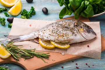 Cooking Raw Dorado Fish with Spinach, rosemary, olives, Herbs, Spices and Lemon closeup on Wooden Cutting Board on an aquamarine background