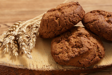 Three pieces of homemade oatmeal cookies and wheat spikes lay on a piece of wood on a brown old table