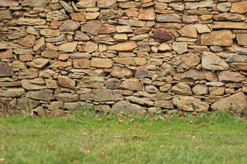 A green grass with autumn leaves in front of a big rustical stone wall with a lot of stones