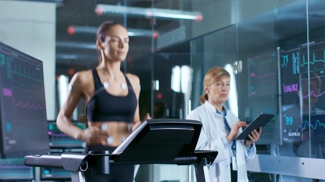 Beautiful Woman Athlete Runs on a Treadmill with Electrodes Attached to Her Body, Female Physician Uses Tablet Computer and Controls EKG Data Showing on Laboratory Monitors.