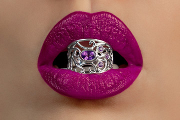 красивые женские губы с сиреневой помадой держат серебреное кольцо с драгоценными камнями © lufotolab