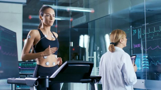 Beautiful Woman Athlete Runs on a Treadmill with Electrodes Attached to Her Body, Female Physician Interacts with Touchscreen and Controls EKG Data Showing on Laboratory Monitors.