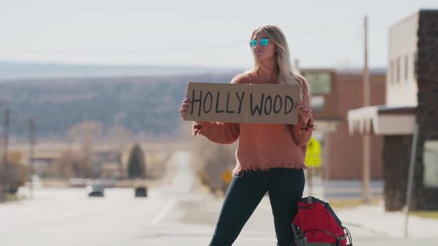 Woman holding Hollywood sign hitchhiking on remote rural road / Loa, Utah, United States