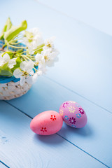 Two painted Easter eggs near basket with white spring flowering branch on light blue background