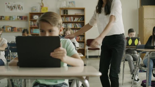 Teacher assisting girl raising hand in classroom reading digital tablet / Provo, Utah, United States