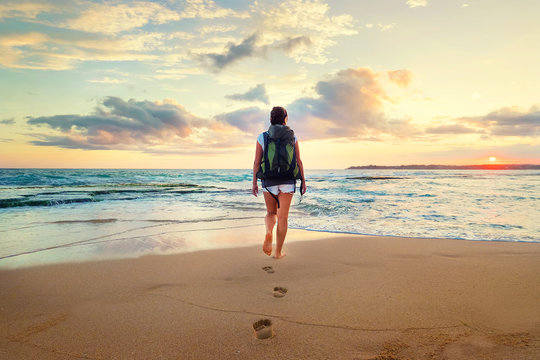 Woman with backpack walk on the ocean sand beach at sunset time