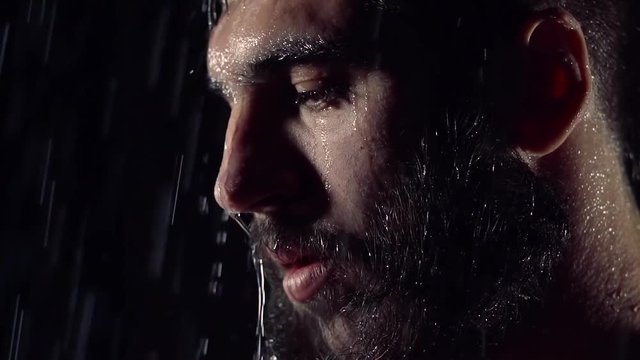 face of handsome bearded man standing under rain in dark street, close-up, looking down and forward