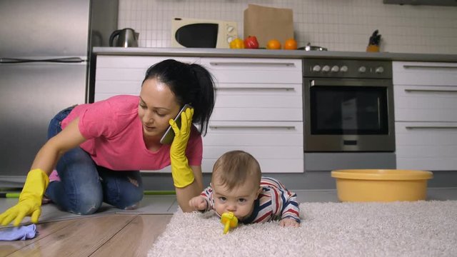 Busy Working Hispanic Mother Managing To Take Care Of Her Baby Son, Clean And Scrub The Floor And Talk Business On Mobile Phone At The Same Time. Young Woman In Rubber Gloves Housekeeping. Dolly Shot