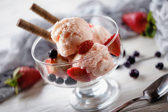 Several scoops of strawberry ice cream with wafer, strawberry and blue berry in glass next to spoon and towel. Summer background.
